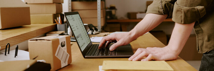 Person organizing tasks at desk, using laptop, cardboard boxes around with various supplies, working environment, engaging in professional activities, workspace filled with materials