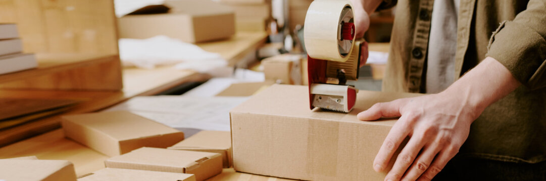 Worker sealing boxes with tape machine in shipping warehouse, preparing packages for delivery, and organizing workspace during warehouse operations