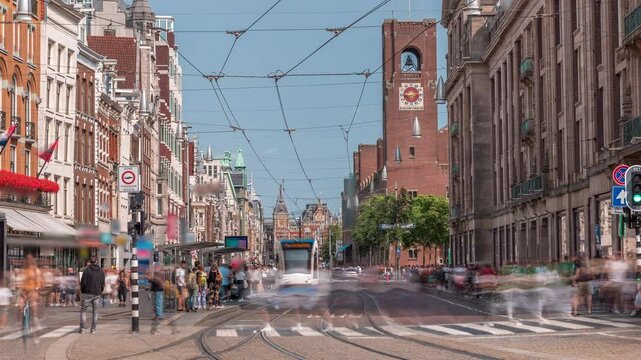 Electric tram on Damrak Street heading from Dam Square to Centraal Station in a timelapse. Traffic moves on the road while people wait at the tram stop. Amsterdam, Netherlands