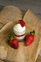 a dessert placed on a wooden board. It appears to be a layered parfait or cheesecake in a small glass, topped with whipped cream and a strawberry. Two whole strawberries are next to the dessert on the