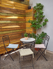 Outdoor seating area with a small round white table surrounded by four metal chairs, and a wooden wall behind. A potted plant in a yellow pot sits at the center of the table.