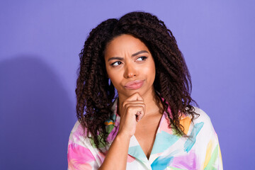 Thoughtful young woman with curly hair pondering against a vibrant purple background, wearing a colorful casual shirt