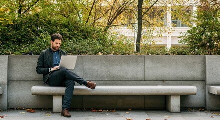 Fototapeta premium Man working on a laptop while sitting on a concrete bench in a park