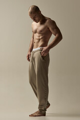 Muscular man, dressed only beige pants, standing barefoot in relaxed pose, gazing down, wearing...