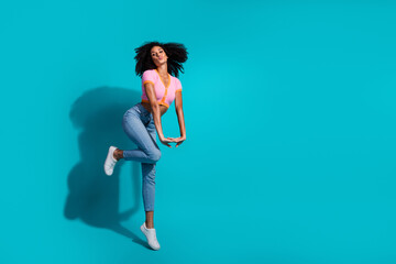 Young stylish woman dancing joyfully against a vibrant turquoise background in casual fashion.