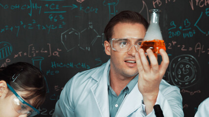 Smart teacher holding solution in beaker while student doing experiment at blackboard with chemical...