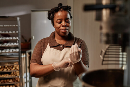 Chest up shot of smiling African American female baker preparing to make pastries putting on vinyl gloves in small bakery kitchen