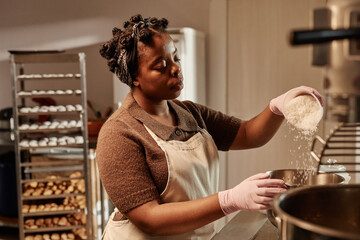 Medium shot of African American female baker pouring coconut flakes from measuring cup mixing ingredients for bread dough in bakehouse kitchen