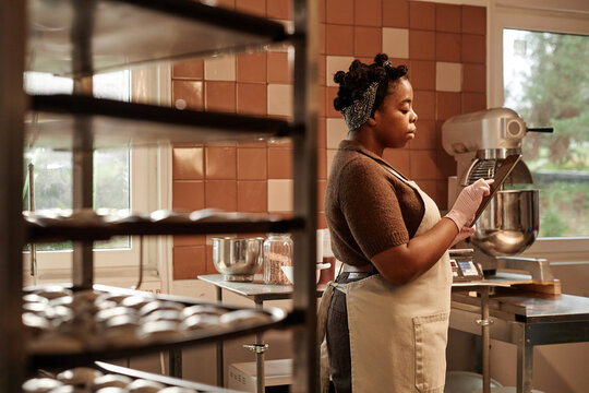 Side view of adult Black female chef wearing apron using digital tablet while cooking in bakery kitchen
