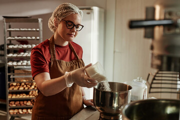 Medium shot of young female bakehouse worker adding shredded coconut into bowl precisely measuring...