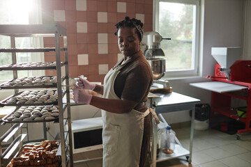 Medium full shot of African American female artisan wearing apron transporting trays of freshly baked rolls placed on mobile rack, during working day in bakery kitchen