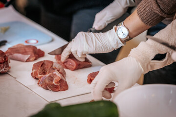 Close-up of hands wearing gloves cutting raw meat on a white cutting board. The scene suggests food preparation, hygiene, and cooking, with a watch visible on one wrist.