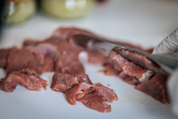 Close-up of hands wearing gloves cutting raw meat on a white cutting board. The scene suggests food preparation, hygiene, and cooking, with a watch visible on one wrist.
