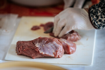 Close-up of hands wearing gloves cutting raw meat on a white cutting board. The scene suggests food preparation, hygiene, and cooking, with a watch visible on one wrist.