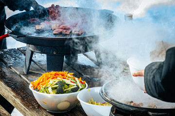 Steaming hot vegetable stir-fry is being cooked outdoors on portable stoves in a snowy setting. Fresh ingredients like zucchini, carrots, and broccoli are prepared in bowls on a rustic table.