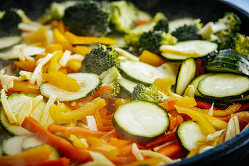 A steaming vegetable stir-fry with zucchini, bell peppers, carrots, and broccoli is being cooked in a pan. A spatula is stirring the fresh, colorful ingredients.