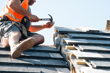 Construction worker installing roofing materials at a residential site while using a hammer and...