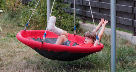 Bored child with leg in plaster lying on garden nest swing