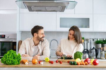 A couple shares cooking tips and laughs in their contemporary kitchen filled with vibrant vegetables and a warm atmosphere.