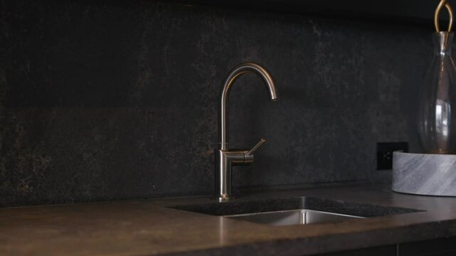Modern kitchen sink with sleek, curved faucet. Countertop and backsplash are made of dark material, possibly granite or similar stone. Decorative glass container on right side