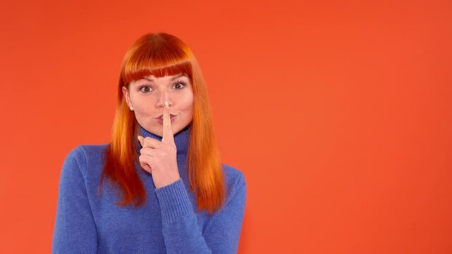 Woman with orange hair making shushing gesture against bright orange background