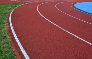 Fototapeta premium Red running track curve with blue sports field in right side and green grass in left side, sunny day