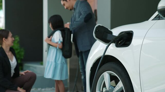 Progressive parents with electric vehicle and home charging station. Happy family with daughter giving each other high fives before leave for school. Alternative future transportation concept of EVs