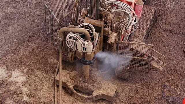 Aerial video of  Heavy construction machinery piling rig dilling into the ground at a construction site rotating and releasing mud, smoke and steam around 