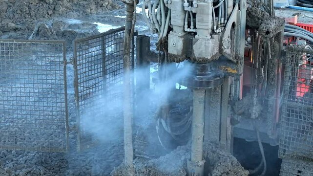 Close-up of Heavy construction machinery piling rig dilling into the ground at a construction site rotating and releasing mud, smoke and steam around 