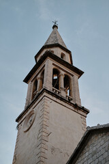 Historic church bell tower with a pointed spire in old town Budva set against a clear blue sky. The aged stone architecture and intricate details reflect timeless elegance and religious heritage.