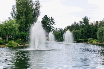 Naklejka premium Ornamental fountain splashing water in a pond surrounded by grass, trees, and flowerbeds in a well-maintained park creates a tranquil atmosphere
