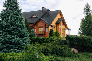 Modern wooden cottage with dark roof and chimneys surrounded by evergreen trees and bushes under cloudy sky in countryside during summer day