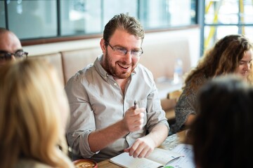 Group meeting in a casual setting with people discussing and smiling. A man with glasses is engaged...
