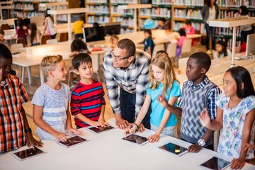 A diverse group of children and a teacher in a library, interacting with tablets. The teacher engages with students, fostering a love for learning and technology. Teacher teaching class in library.