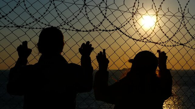 Refugees looking at sunset behind barbed wire fence. Silhouetted refugees gazing through barbed wire fence, embodying hope and longing for freedom amid harsh border conditions