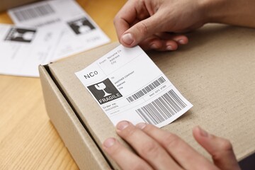 Naklejka premium Man sticking shipping label with barcode on parcel at wooden table indoors, closeup