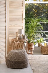 Stylish knitted pouf, wicker furniture and beautiful houseplants in room. Interior design