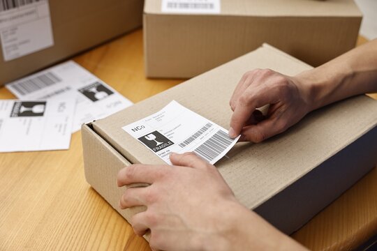 Man sticking shipping label with barcode on parcel at wooden table indoors, closeup