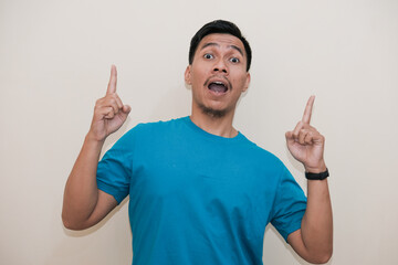 Fototapeta premium A Southeast Asian man in a turquoise green tshirt smiling and pointing up with both index fingers, in a white background. The image captures an exciting and curious expression 