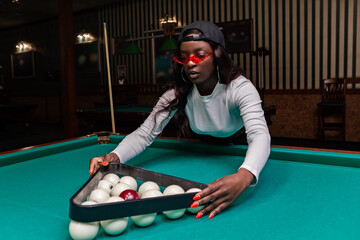 Young woman wearing stylish red sunglasses and a backwards baseball cap leans over a billiard...