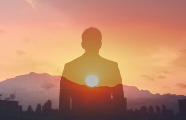 A businessman looks out over the city skyline at sunset visualizing success. Man silhouette facing New York City, people reaching goals, having a vision, never give up concept 
