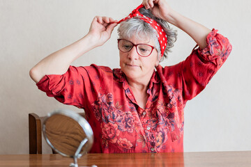 Senior woman in red adjusting headscarf with mirror on wooden table