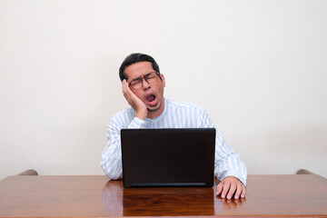A man yawning showing tiredness when sitting in front of a laptop