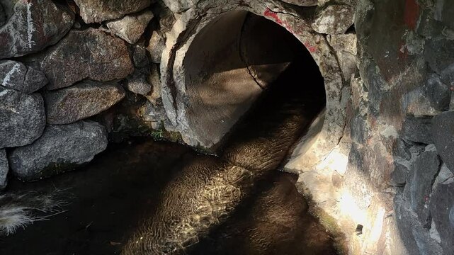 Old underground drainage collector pipe comes to the surface with sunlight reflections from water on the pipe and stone walls.
