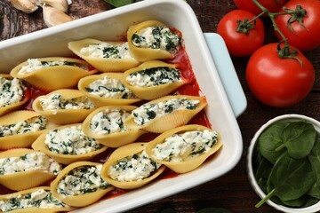 Delicious conchiglie pasta with ricotta cheese, spinach and tomato sauce in baking dish on wooden table, flat lay