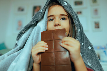 Little Girl Hiding under a Blanket Eating a Chocolate. A small child concealed in her room tasting...