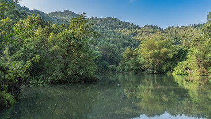 Beautiful calm lake. There are thickets of lush green trees on the banks.  Mountains against a clear blue sky. Reflection on the water. China. Tianxingqiao Scenic Area. Guizhou