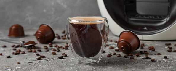 Glass of delicious espresso, capsule coffee maker, pods and beans on grey grunge table