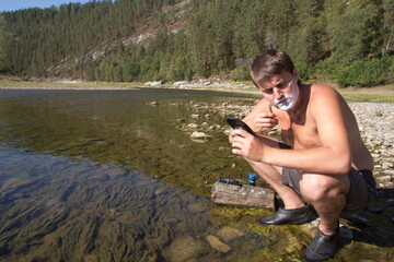 young handsome man shaving by the river white against the backdrop of the Ural Mountains on a summer sunny morning