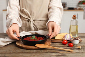 Young woman with frying pan of tomato paste and ingredients on wooden table in kitchen